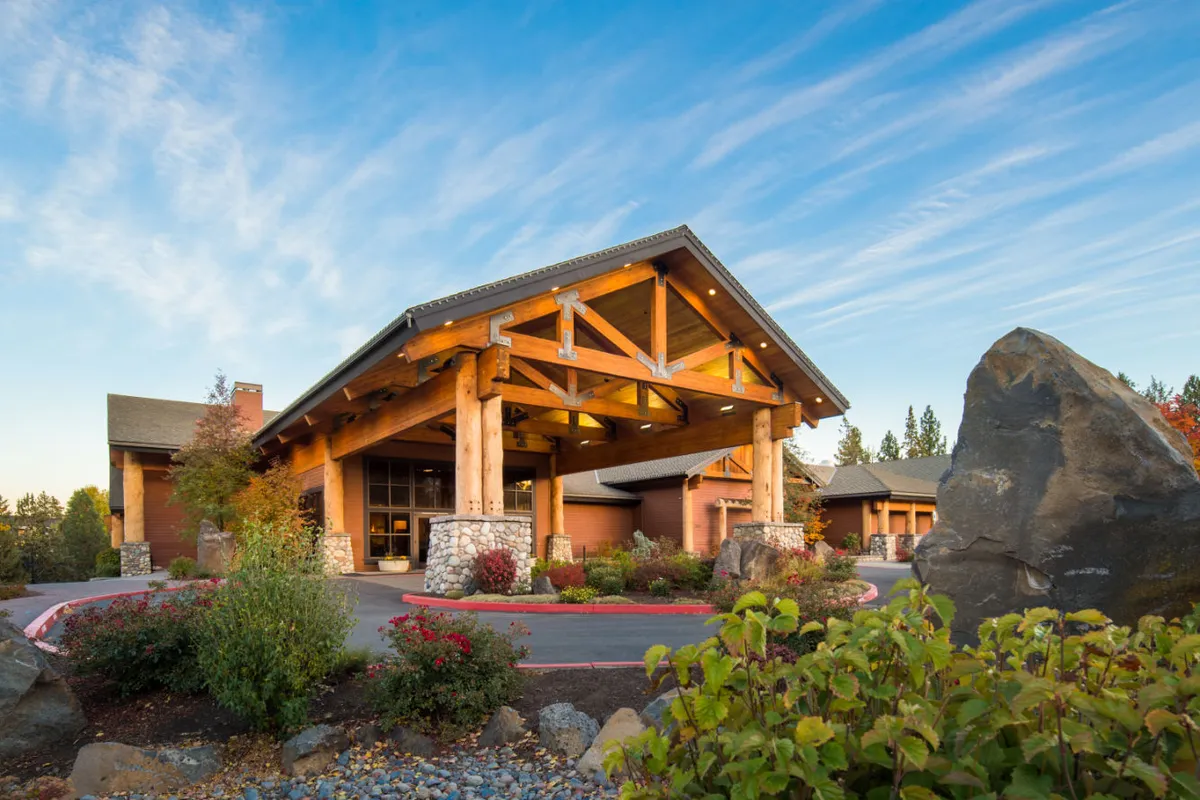This exterior image captures the main entrance of Riverhouse Lodge, adorned with large wooden beams and surrounded by beautifully landscaped gardens and large boulders. The setting sun adds a warm, romantic glow, and the natural elements create a tranquil atmosphere that hints at the scenic riverfront location.