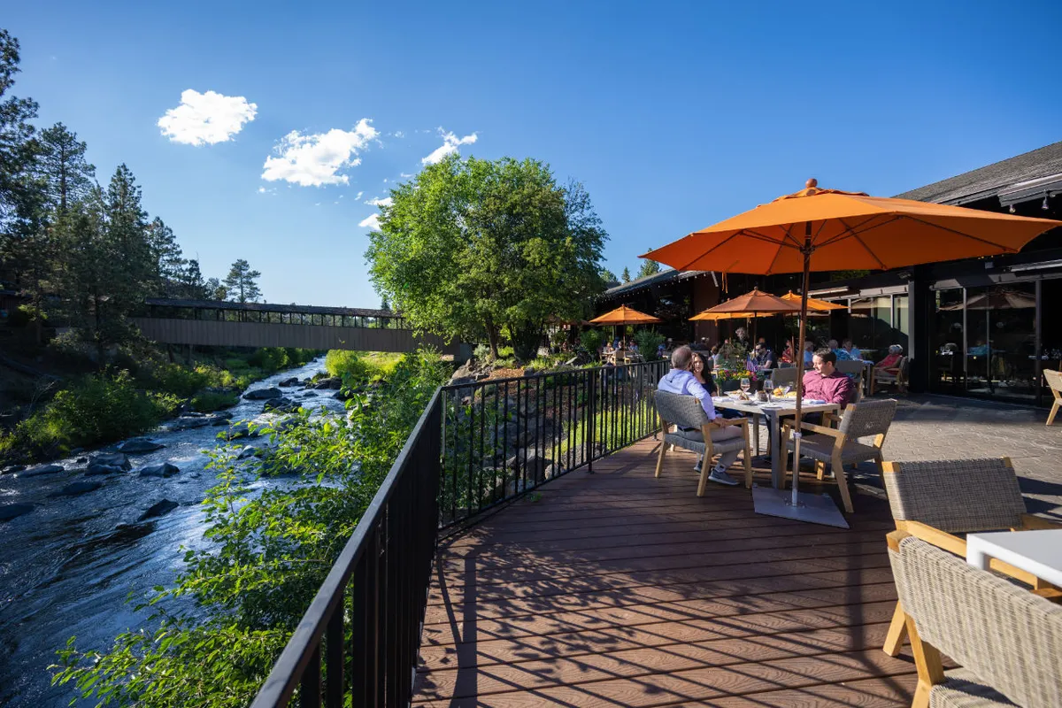 An exterior shot of the river deck adjacent to the lounge, with guests dining and socializing on a patio shaded by orange umbrellas. The fire pit glows as the sun sets over the Deschutes River, framing a scene of relaxed alfresco enjoyment.