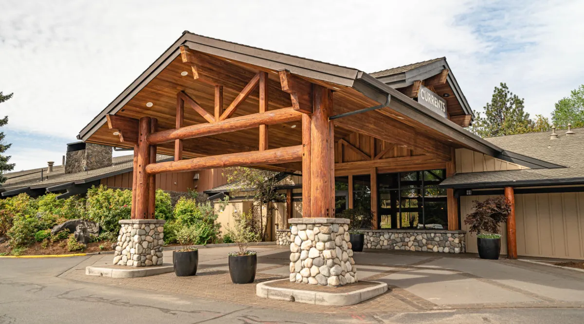 The image showcases the Riverhouse Lodge's glass entrance framed by large wooden beams and stone pillars. Lush landscaping surrounds the rustic yet inviting entrance, hinting at the welcoming hospitality guests receive upon arrival. The scene captures guests arriving and interacting in the entrance area, reinforcing a sense of seamless service and ease.