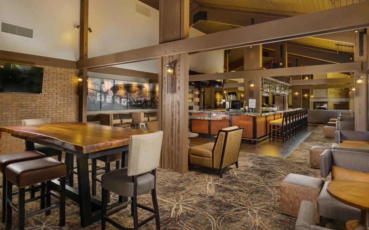 A welcoming interior scene of the CURRENTS lounge showcasing a stylish wooden bar, a large communal table, comfortable seating areas, and backlit bar shelves. Several TVs are visible, and guests are seen socializing, enjoying drinks, and playing games.