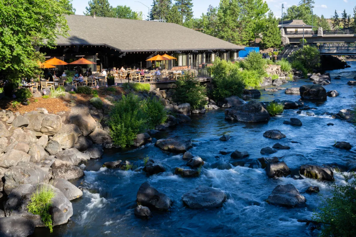 The image showcases an inviting restaurant patio set against the backdrop of the flowing Deschutes River, lush greenery, and dramatic rock formations. The scene is bathed in the warm hues of sunset, suggesting a tranquil and picturesque setting perfect for outdoor dining or quiet relaxation by the water. Wildlife sightings and hints of seasonal events like outdoor concerts add to the lively yet peaceful ambiance.
