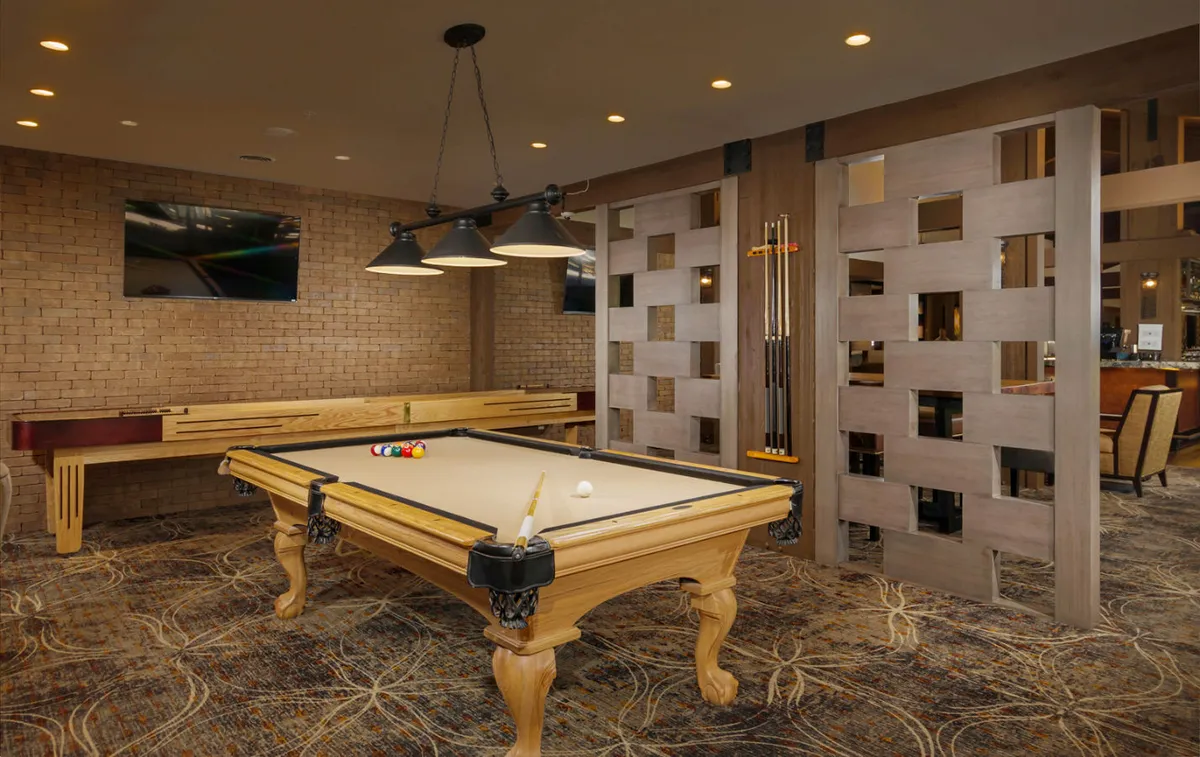 An inviting interior shot of a game area within the lounge, featuring a pool table under modern lighting fixtures and a decorative screen in the background. Guests are seen playing pool and socializing nearby, with TVs present for sports viewing.