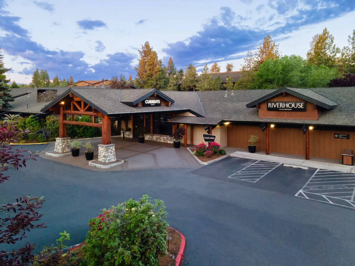 A wider exterior perspective of the main entrance, surrounded by lush landscape greenery, with prominent signage for both CURRANTS and RIVERHOUSE. The scene implies opportunities for outdoor dining, relaxation in the gardens, and attending cultural or culinary events, reinforcing the venue's role as a hub for both work-related gatherings and casual socializing.