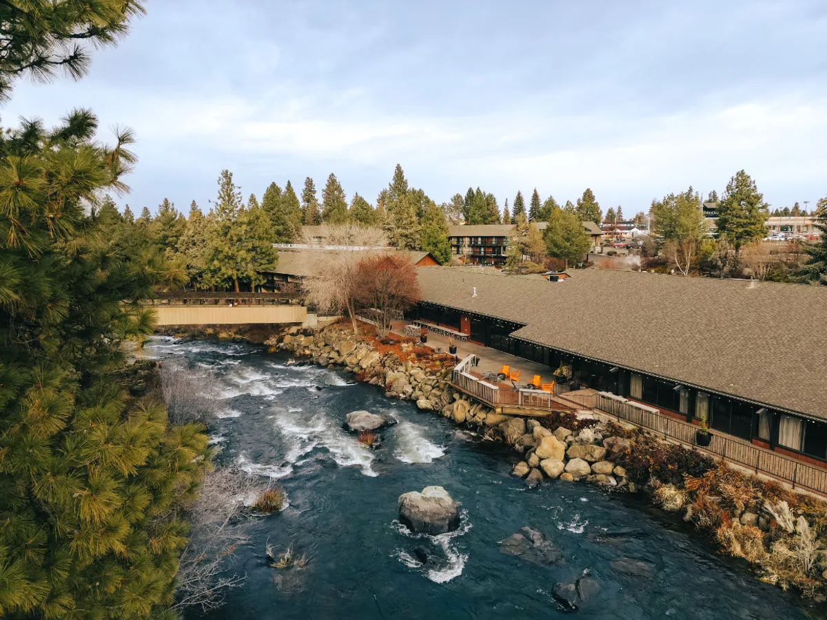 A tranquil scene showing a riverside building with a deck area overlooking the water, surrounded by trees and lush greenery. The outdoor space connects the indoors to the natural beauty of Central Oregon, highlighting opportunities for relaxation and scenic views.