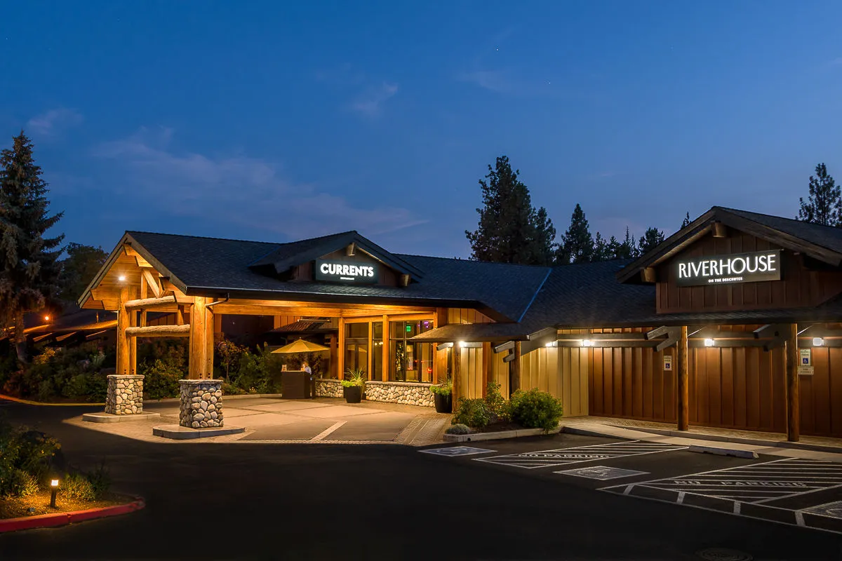 The exterior entrance canopy of Riverhouse Lodge is prominently displayed, with clear signage for both Currents and Riverhouse, and a thoughtfully landscaped area framing the view. The scene is set at sunset, enhancing the welcoming and professional atmosphere. There are hints of people gathering for dining or social events, suggesting a lively yet relaxed setting.